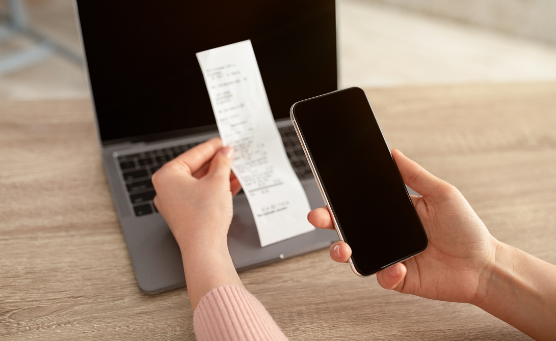 persoon maakt foto van kassabon met telefoon met laptop op de achtergrond